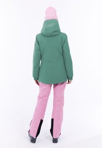 Chaqueta impermeable verde con capucha, combinada con pantalones de esquí rosas con acentos negros. Textura suave, diseñada para actividades invernales.