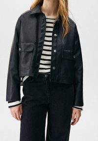 Femme portant une veste en cuir noire courte boutonnée sur une chemise à manches longues rayée noir et blanc et un pantalon noir taille haute.