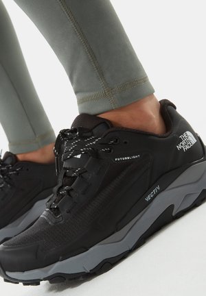 Zwarte wandelschoenen met een bovenwerk van gestructureerd mesh, dikke grijze tussenzool, zwarte rubberen buitenzool en reflecterende veters, gecombineerd met groene legging.