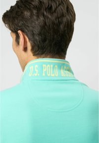 Turkoosi pikeepaita, jossa on keltainen "U.S. POLO ASSN." -logo kauluksessa, yksinkertainen muotoilu ja pehmeä rakenne.