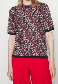 Kurzarmpullover in Schwarz mit einem bunten Blumenmuster, geripptem Kragen und Bündchen, kombiniert mit roten Hosen.