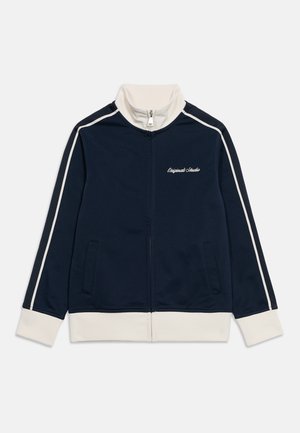 Sötétkék és fehér cipzáras tréningdzseki magas gallérral, fehér csíkokkal az ujjakon, elülső zsebekkel és "Originals Studio" felirattal a mellkason.