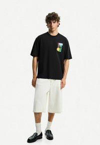 Black short-sleeve T-shirt featuring a colorful graphic design on the left chest, paired with white knee-length shorts and black loafers.