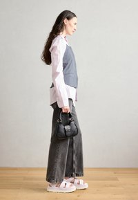 Harmaa liivi vaaleanpunaisen paidan päällä, löysät harmaat farkut ja valkoiset korokepohjasandaalit. Musta käsilaukku, jossa on soljet yksityiskohdat, pidetään kädessä.