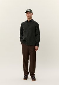 Άνδρας που φοράει σκούρο πουκάμισο με λεπτές ρίγες, καφέ παντελόνι, σκούρα παπούτσια και πράσινο καπέλο μπέιζμπολ, στέκεται με το ένα χέρι στην τσέπη.