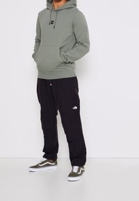 Personne portant un sweat à capuche gris-vert, un pantalon noir North Face coupe décontractée et des baskets Vans vertes, debout avec les mains dans la poche du sweat.