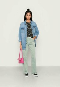 Casaco de ganga azul claro por cima de uma camisa preta com flores, combinado com calças verde menta e ténis pretos. A modelo segura uma bolsa pequena rosa.