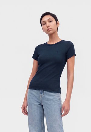 Femme aux cheveux courts noirs porte un t-shirt bleu foncé ajusté à manches courtes et un jean bleu clair, debout devant un fond clair uni.