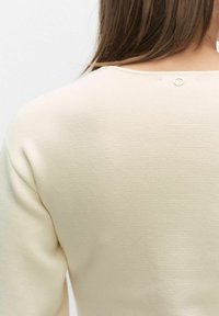 Roomkleurige gebreide top met een gestructureerd oppervlak en een ronde halslijn, met een klein rond accent vlak bij de achterkant van de nek.