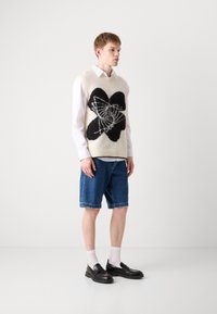 Gilet en laine tricoté, crème avec un grand motif abstrait noir, porté sur une chemise blanche. Assorti avec un short en jean et des mocassins noirs.