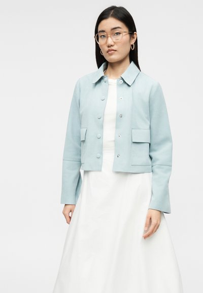 Γυναίκα με μακριά μαύρα μαλλιά, φορώντας στρογγυλά γυαλιά, χρυσούς κρίκους σκουλαρίκια, ανοιχτό μπλε σακάκι με κουμπιά και λευκό φόρεμα, στέκεται μπροστά σε απλό φόντο.