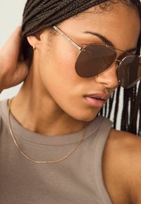 Bruine pilotenzonnebril met een dunne metalen rand, gedragen met een beige geribde tanktop en een delicate gouden ketting.