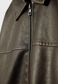 Bruine leren jas met een ritssluiting, voorzien van een kraag en een soepele textuur. Opvallende stikseldetails zijn zichtbaar langs de naden.