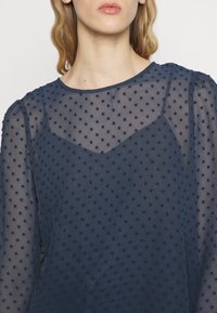 Blusa azul-marinho com padrão de bolinhas, tecido semi-transparente, decote redondo e mangas longas bufantes com detalhes em camada.