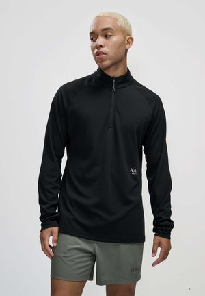Sort langærmet træningsbluse med kvart lynlås-design, tekstureret stof og logo-detaljer, parret med grønne shorts.