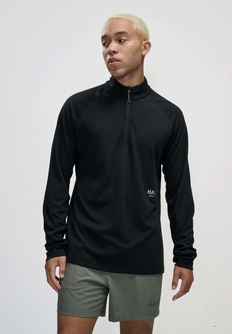 Schwarzer Pullover mit langen Ärmeln, einem Viertel-Reißverschluss, strukturiertem Stoff und einem kleinen Logo auf der Vorderseite. Kombiniert mit olivgrünen Shorts.