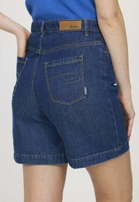 Shorts en denim de couleur bleu foncé, avec des poches arrière et une étiquette en cuir marron au niveau de la taille. Ourlet propre et détails de couture subtils.