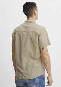 Camisa de manga corta en color beige con un corte relajado, cuello y detalles de costura visibles. Combinada con jeans azules de tela texturizada.