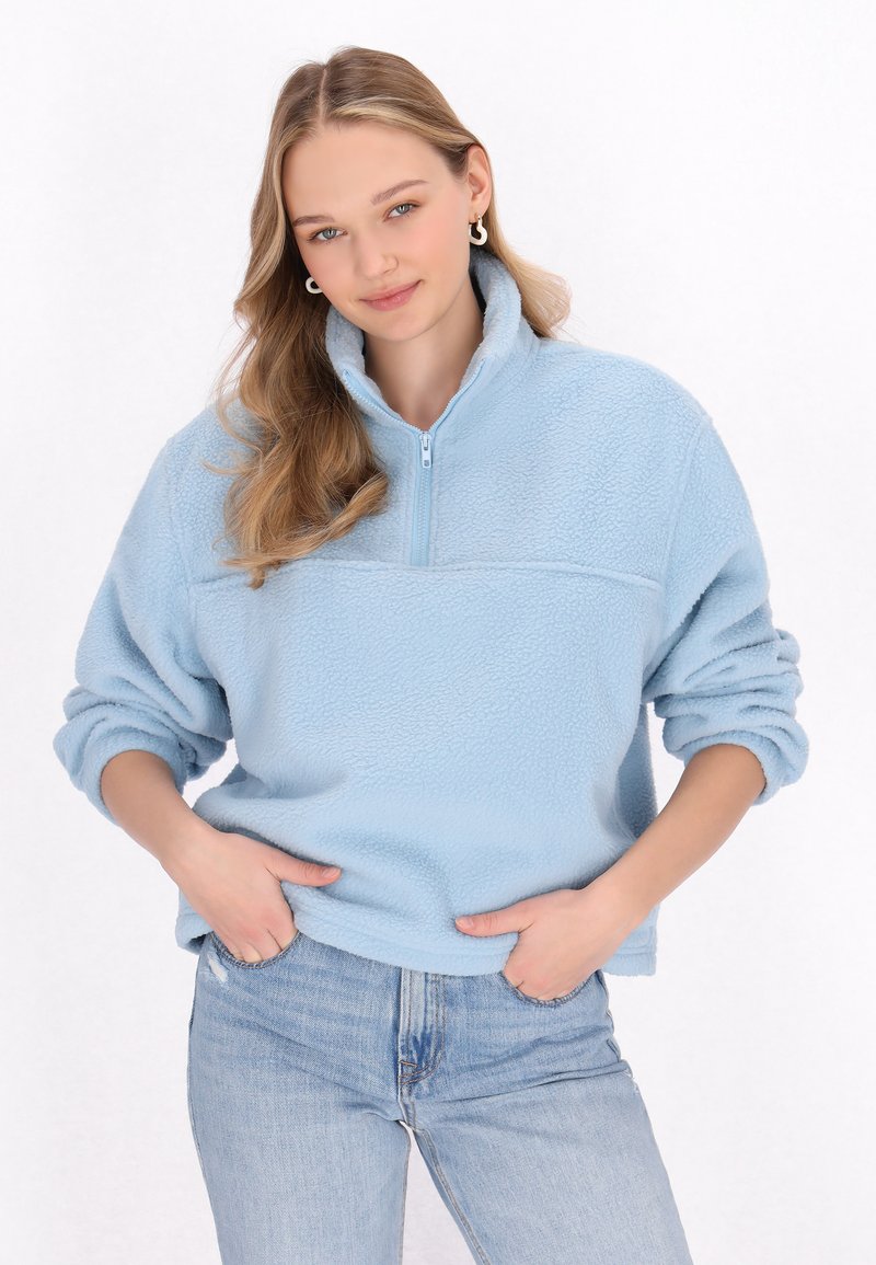 Pullover in pile azzurro chiaro con collo a zip quarti, caratterizzato da un tessuto testurizzato, maniche lunghe e una vestibilità comoda. Abbinato a jeans in denim chiaro.