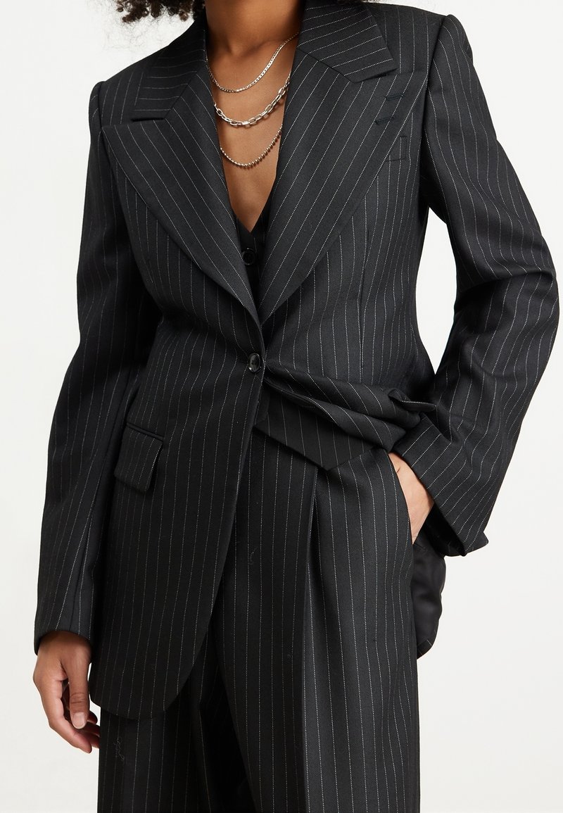 Czarna marynarka w cienkie prążki z szerokimi klapami, pojedynczym zapięciem na guzik i przednią kieszenią. Tkanina o fakturze z dodatkiem metalicznych nici.