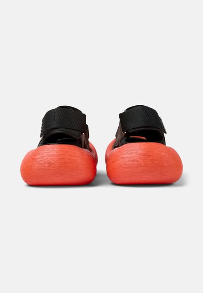 Sandales noires avec larges brides et semelles épaisses texturées de couleur orange vif, vues de dos sur un fond blanc.