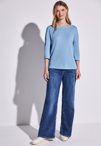Helles blauer, langärmeliger Pullover kombiniert mit dunklen, ausgestellt geschnittenen Jeans und weißen Sneakers; glatte Textur, lockere Passform, minimalistisches Design.