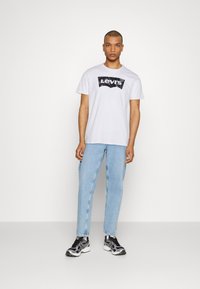 Hvit bomull t-skjorte med svart Levi's logo grafikk, kombinert med lyse blå denimjeans og svart-hvite treningssko.