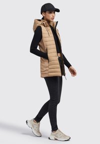 Gilet imbottito beige con cappuccio su una maglia nera a maniche lunghe in coste, abbinato a jeans skinny neri e sneakers beige con dettagli blu.