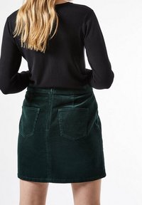 Grön kordflors mini kjol med två bakfickor och en slät yta. I kombination med en svart långärmad topp, sedd från baksidan.