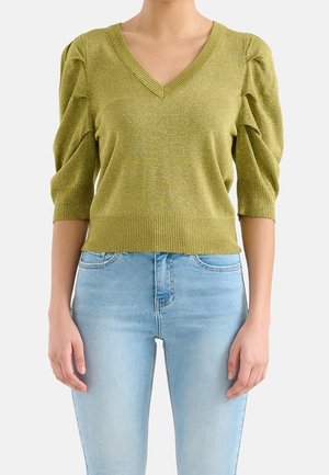Femme portant un pull vert clair à col en V avec des manches bouffantes jusqu'au coude et un jean taille haute bleu clair sur fond blanc.