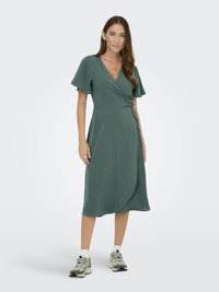 Abito avvolgente verde con maniche a sbuffo corte, una cintura legata in vita e un orlo fino al polpaccio. Indossato con sneakers sportive e calzini alla caviglia.