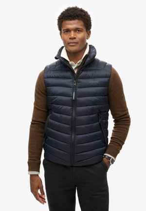 Chaleco tipo puffer azul marino con diseño acolchado, cierre delantero con cremallera, cuello alto y dos bolsillos laterales, llevado sobre un suéter marrón. Reloj plateado en la muñeca.