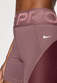 Σορτς Nike Pro σε απαλό μωβ χρώμα με ευρεία ζώνη μέσης που φέρει τη λέξη "PRO" σε ανοιχτό ροζ. Ομαλή υφή με πάνελ από δίχτυ στο πλάι.