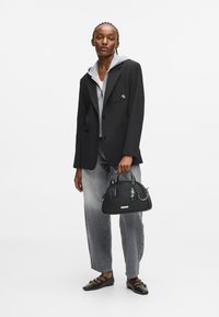 Γυναίκα με μαύρο σακάκι πάνω από γκρι φούτερ και τζιν, κρατώντας μια μαύρη τσάντα με γοητείες, φορώντας μαύρες �μυτερές φλατ με λουράκια.