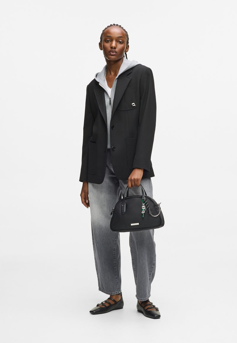 Γυναίκα με μαύρο σακάκι πάνω από γκρι φούτερ και τζιν, κρατώντας μια μαύρη τσάντα με γοητείες, φορώντας μαύρες μυτερές φλατ με λουράκια.