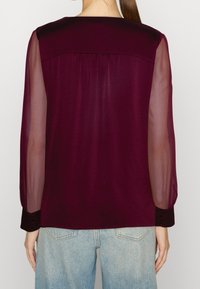 Blusa a maniche lunghe con retro in raso bordeaux, maniche trasparenti e polsini abbottonati, con vestibilità rilassata e dettaglio di piega sul retro.