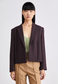 Blazer recortado de color púrpura oscuro con una textura suave, con solapas en pico y mangas largas, combinado con una blusa verde clara y pantalones beige.