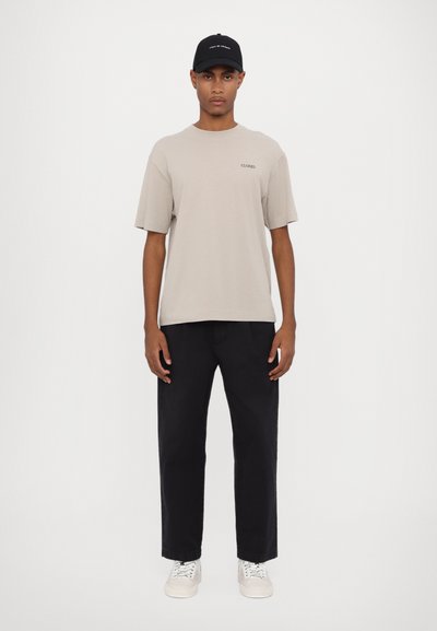 Beige bomull t-skjorte med kort erm og en liten logo på brystet, kombinert med svarte løstsittende bukser og hvite sneakers. Svart caps.