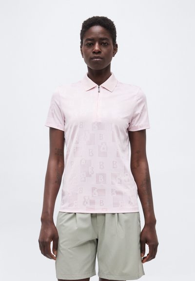 Personne debout, tournée vers l'avant, portant un polo à manches courtes rose clair avec fermeture éclair et un short plissé gris clair, sur un fond uni.