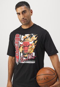 Schwarzes Baumwoll-T-Shirt mit einem Chicago Bulls Grafikdesign, das Flammen, fetten roten Buchstaben und einem Basketball-Icon enthält. Der Text umfasst Team- und Divisionsdetails.
