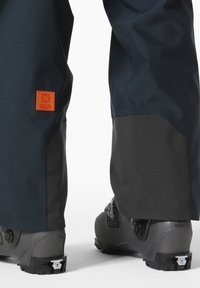 Nepromokavé lyžařské kalhoty v tmavě modré barvě s černými spodními panely, s výraznou oranžovou nášivkou. Odolný materiál a zesílené lemy na kotnících.