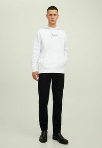 Weißes Hoodie mit Logo, Fronttasche und verstellbaren Kordeln, kombiniert mit schwarzen Hosen und schwarzen Stiefeln. Einheitlicher Hintergrund.