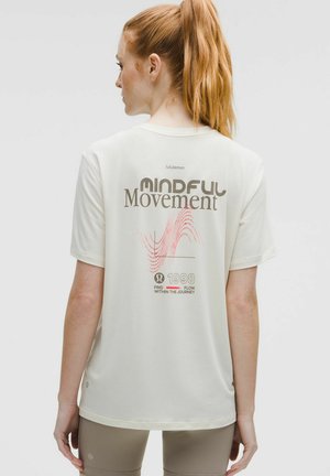 Vrouw met rood haar in een staart, gekleed in een crèmekleurige Lululemon T-shirt met de tekst "Mindful Movement" en beige leggings, met de rug naar ons gekeerd.