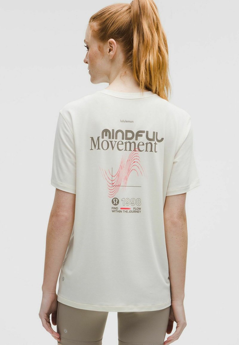 Frau mit roten Haaren im Pferdeschwanz, die ein cremefarbenes Lululemon T-Shirt mit dem Aufdruck "Achtsame Bewegung" und beigefarbenen Leggings trägt, mit dem Rücken zur Kamera.