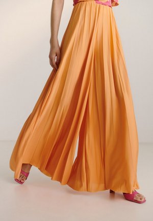 Pantalon orange fluide à jambes larges et plissées, porté avec des talons ouverts roses, montrant le mouvement et la texture du tissu sur un fond uni.