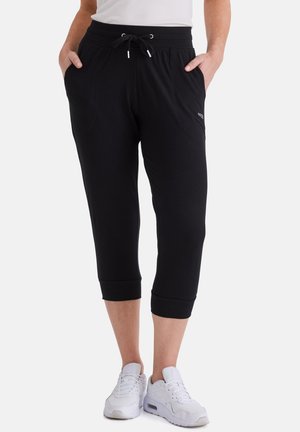 Person trägt schwarze kurze Jogginghosen mit Seitentaschen und weißen Sneakers, stehend vor einem einfarbigen hellen Hintergrund.