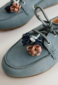 Mocassins en cuir bleu avec semelles en caoutchouc marron, ornés de pampilles en cuir multicolores et de fermetures à lacets sur un fond neutre.