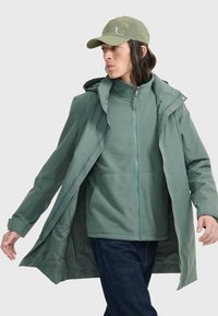 Veste imperméable verte avec capuche, superposée à un zip polaire assorti. Comprend des poches et une fermeture éclair contrastée. Portée avec une casquette.