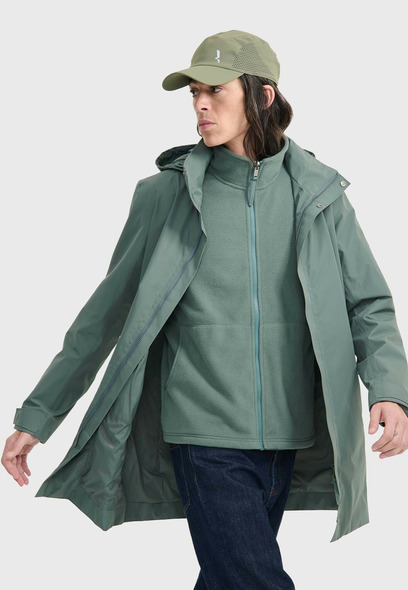 Veste imperméable verte avec capuche, superposée à un zip polaire assorti. Comprend des poches et une fermeture éclair contrastée. Portée avec une casquette.