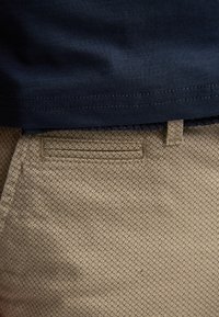 Gros plan sur un pantalon beige texturé et un ourlet de chemise bleu marine, montrant les détails de la poche et des passants de ceinture.